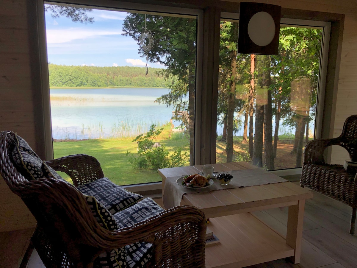 A serene seating area is depicted with woven chairs positioned by large windows. The view showcases a calm lake surrounded by trees, with greenery visible outside. A low wooden table is present, adorned with plates of food, inviting relaxation and connection with nature.
