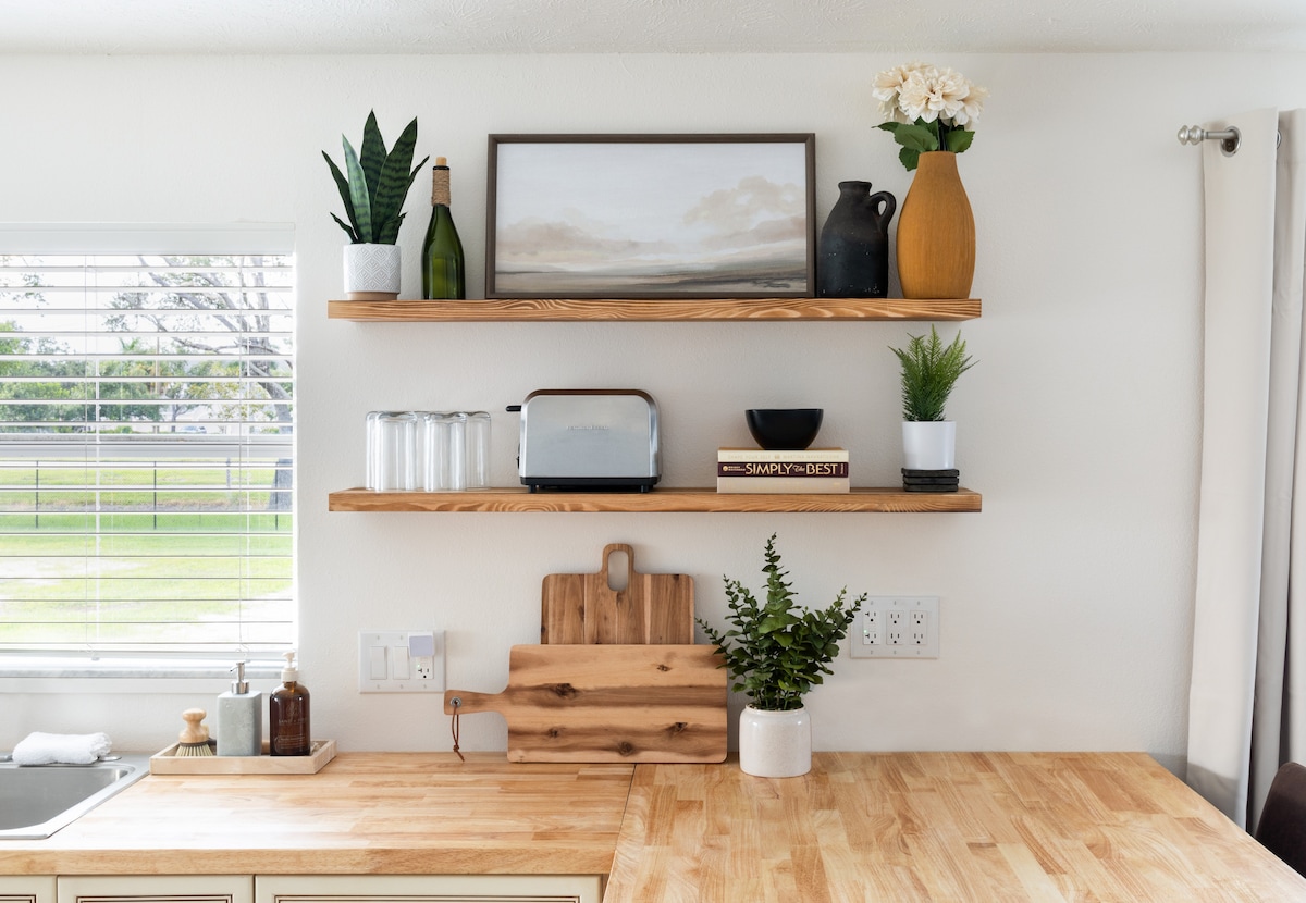 Open shelving is displayed above a wooden countertop, featuring potted plants, a framed painting, a wine bottle, and various decorative items. A silver toaster and glassware are organized neatly, contributing to a modern kitchen aesthetic. Natural light filters through a nearby window.