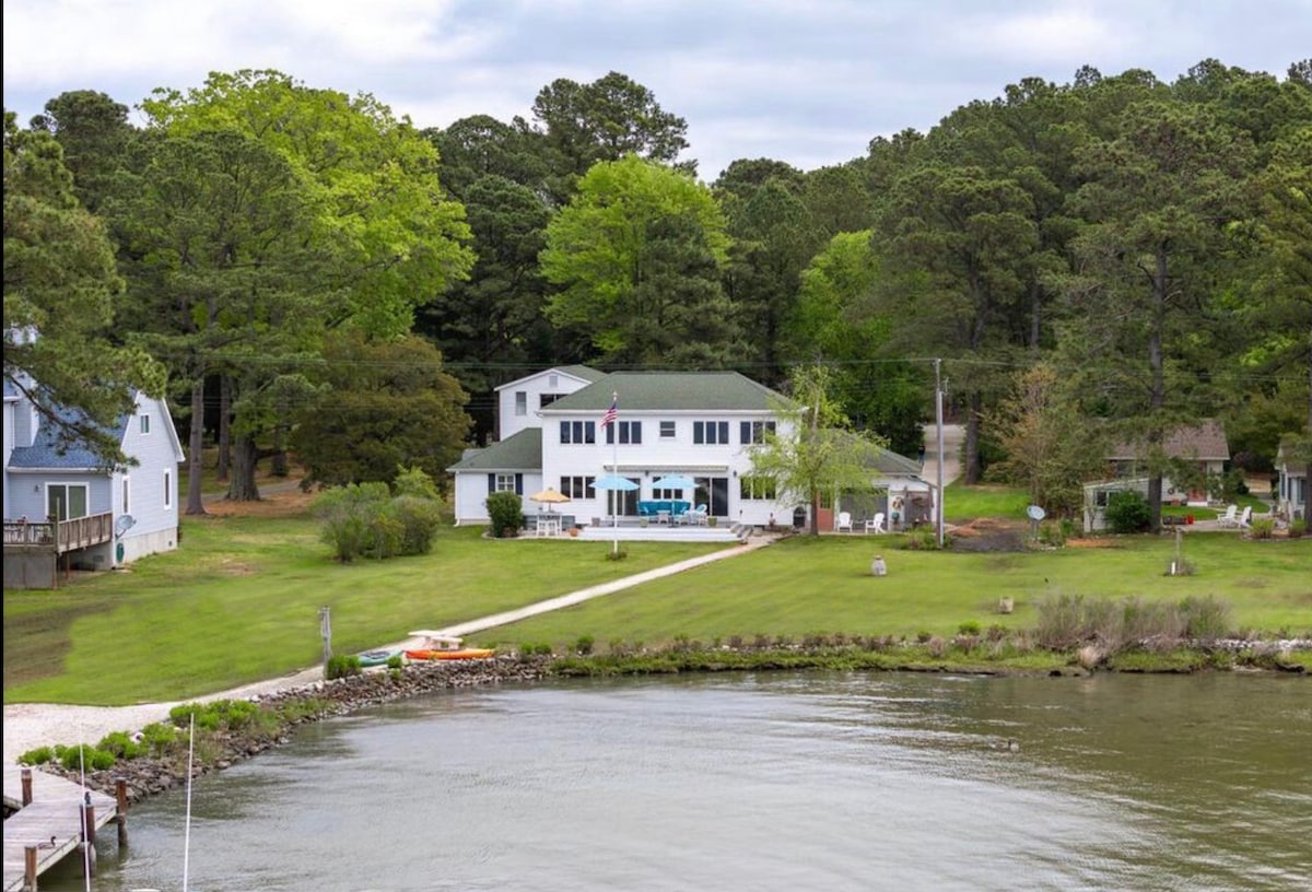 A well-maintained home is situated on the edge of a peaceful cove, surrounded by lush greenery. The spacious yard features a clear path leading to the water, with kayaks visible near the shoreline. The backdrop is marked by trees and a cloudy sky.