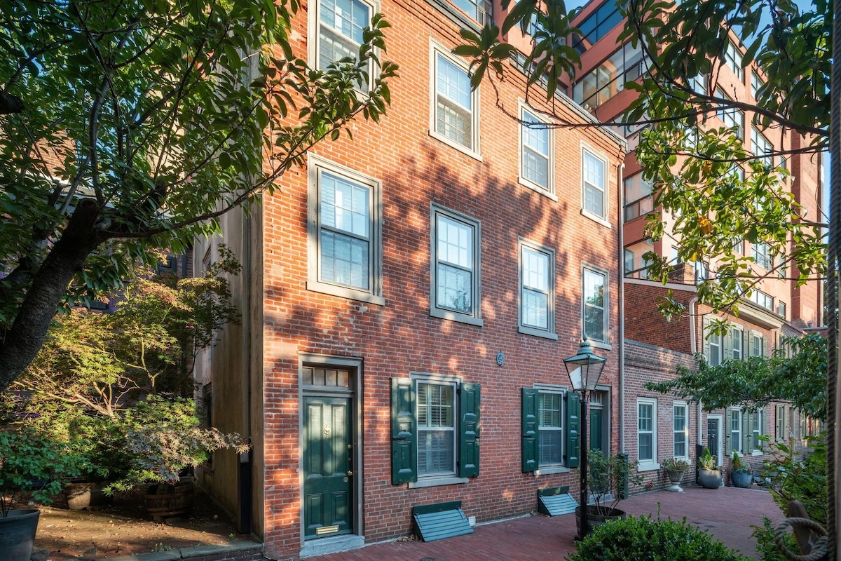 A historic brick building features multiple windows outlined in green shutters, surrounded by lush greenery and a tranquil courtyard. A classic lamp post adds a charming detail to the outdoor space, which is softly illuminated by the warm light of the setting sun.