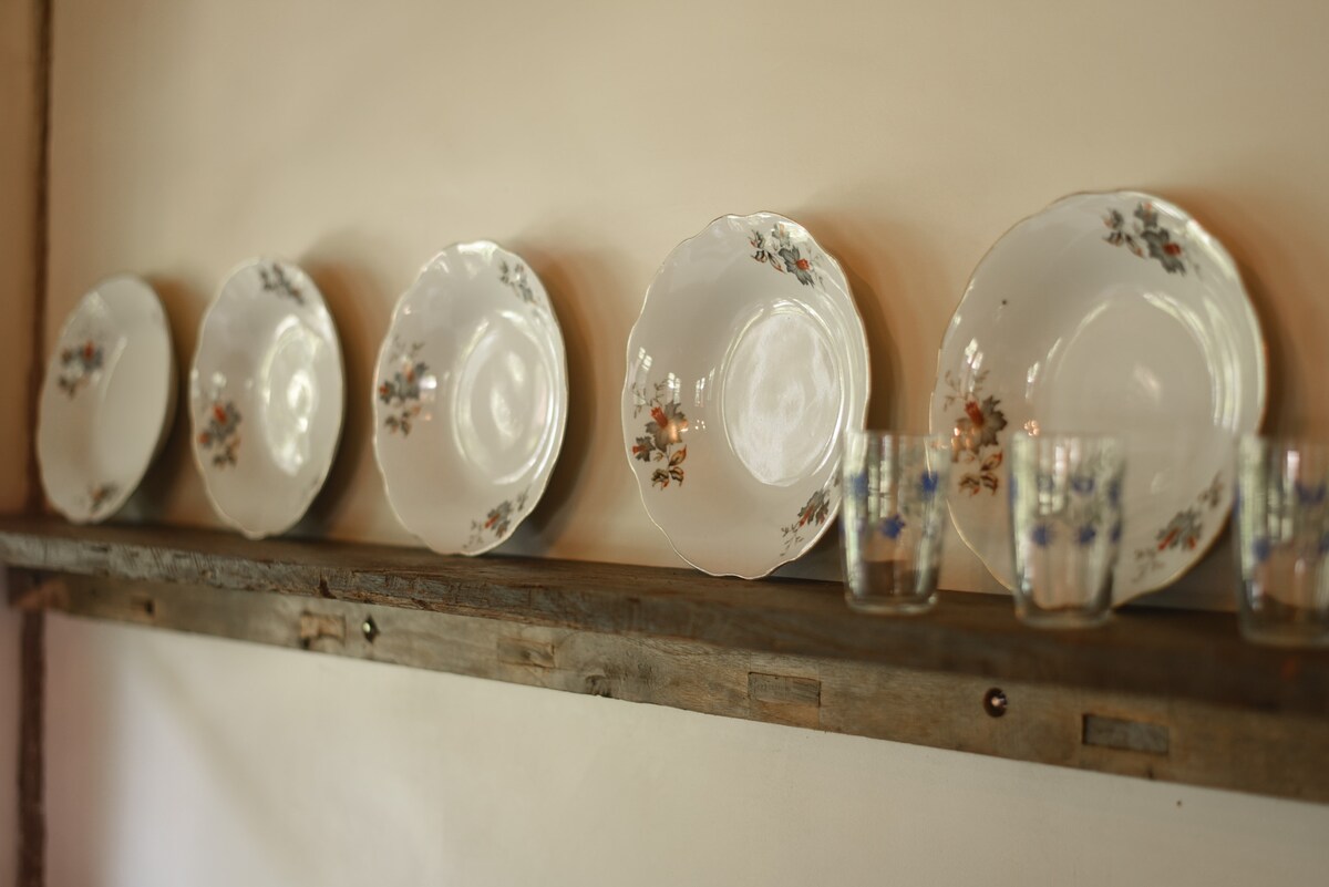 A wooden shelf displays a set of five decorative plates and several glasses. The plates are adorned with floral patterns, while the glasses feature a simple design. The warm tones of the shelf complement the charming arrangement.