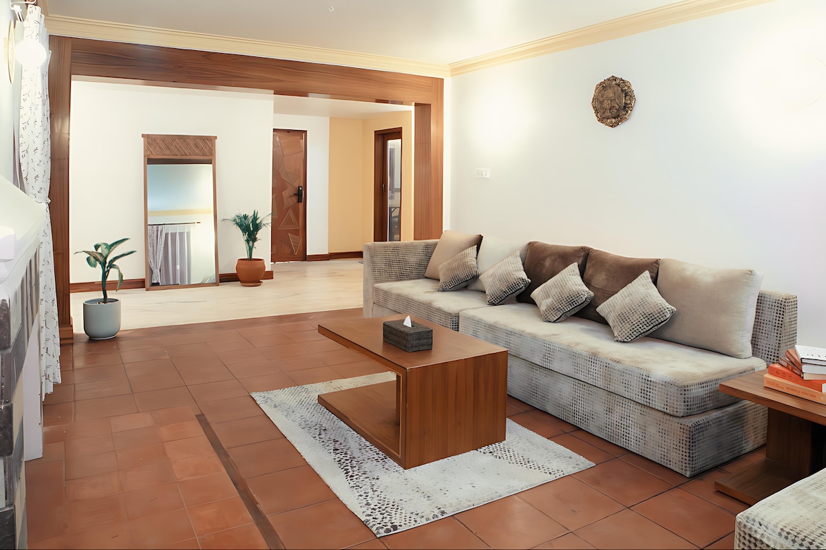 An inviting living area features a spacious sectional sofa adorned with plush pillows. A wooden coffee table sits atop a soft area rug. Natural light enters through large windows, and a decorative plant adds a touch of greenery to the space.
