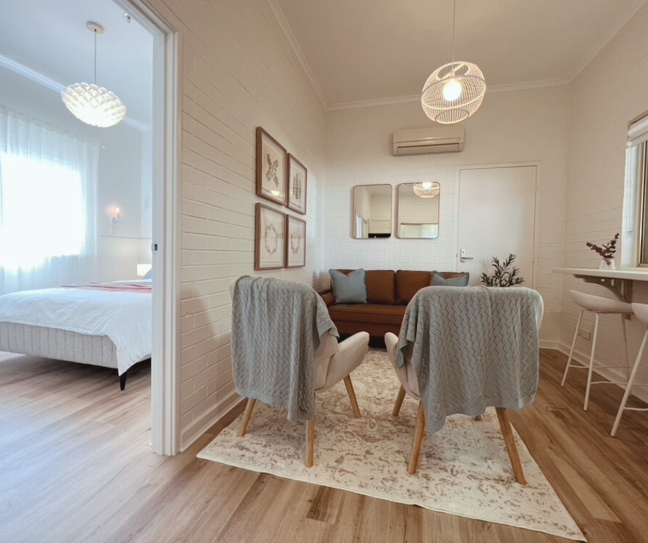 An open-plan living area showcases two light-toned armchairs draped with soft blankets, positioned on a stylish area rug. A comfortable sofa is situated against the wall, complemented by a mirror trio above it. The spacious bedroom area is visible in the background.