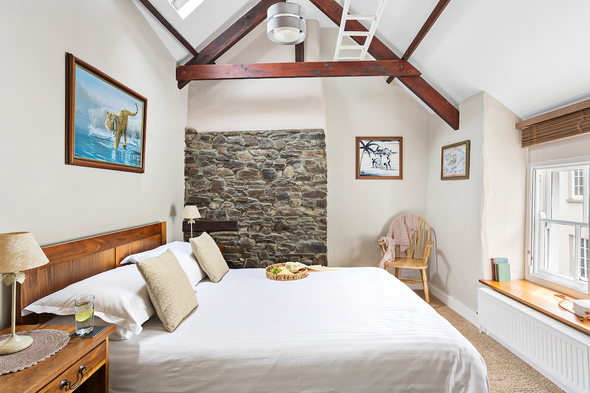 A cozy bedroom features a spacious bed dressed in white linens, complemented by soft pillows. Exposed wooden beams and a stone accent wall add character. A small sitting area with a chair and a few wall art pieces enhance the inviting environment.