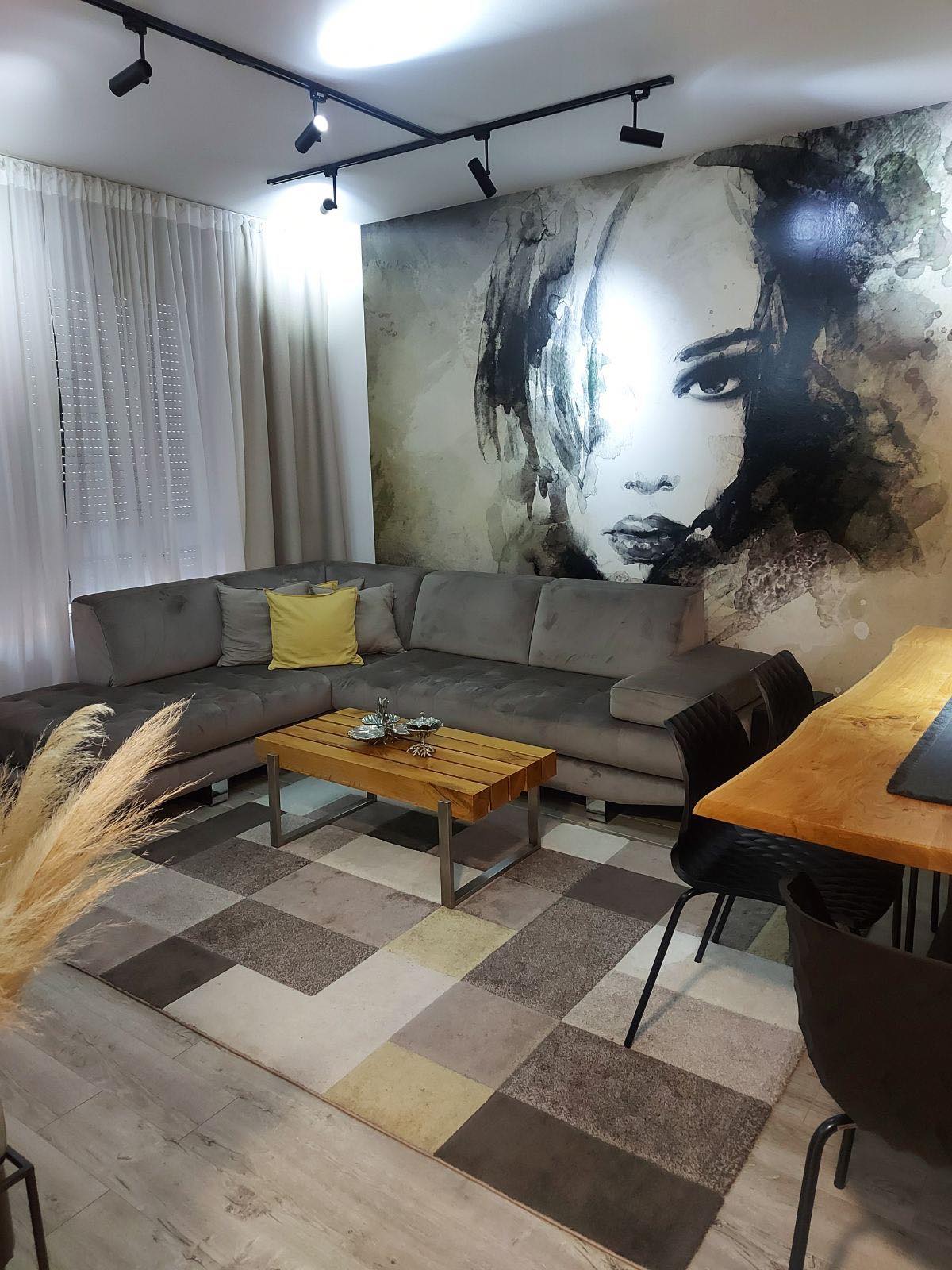 A modern living space features a comfortable sectional sofa in gray and a wooden coffee table. An artistic mural depicting a portrait adds intrigue to the room. Soft curtains allow natural light to filter in, and a patterned rug adds texture to the hardwood floor.