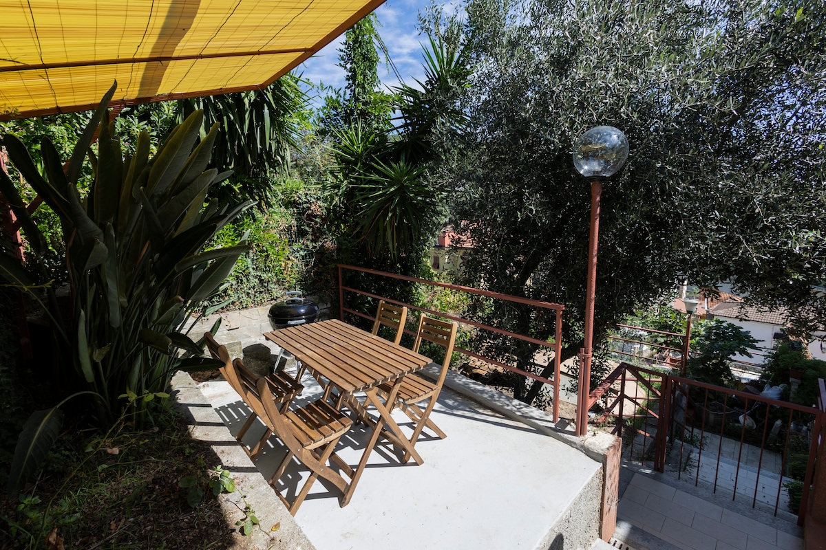 An outdoor terrace is presented, featuring a wooden table surrounded by six matching chairs. Lush greenery, including trees and shrubs, encircles the area, while a protective awning provides shade. A lamp stands nearby, illuminating the space for evening use.