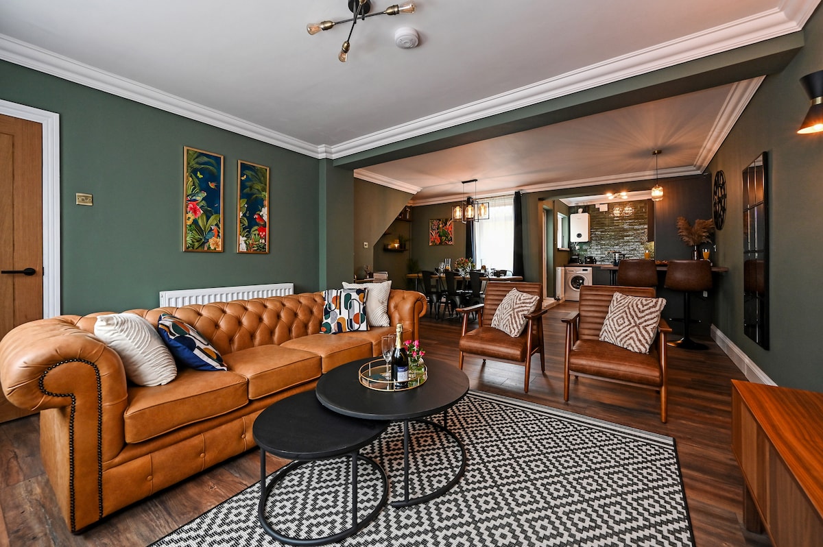 An open-plan living area features dark green walls and a spacious tan leather sofa paired with stylish brown armchairs. A geometric rug anchors the seating area, while a round coffee table sits in the center. The dining area seamlessly connects, showcasing a modern design.