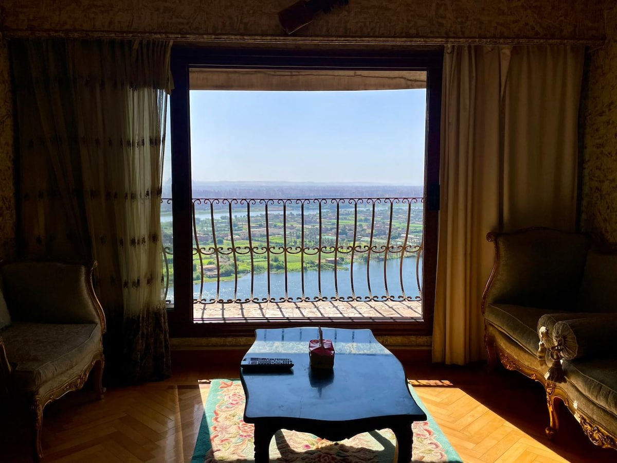 A spacious living area features a large window overlooking the vibrant Nile and lush landscape. Elegant armchairs are positioned near a decorative coffee table. Soft, natural light filters through sheer curtains, enhancing the serene atmosphere.