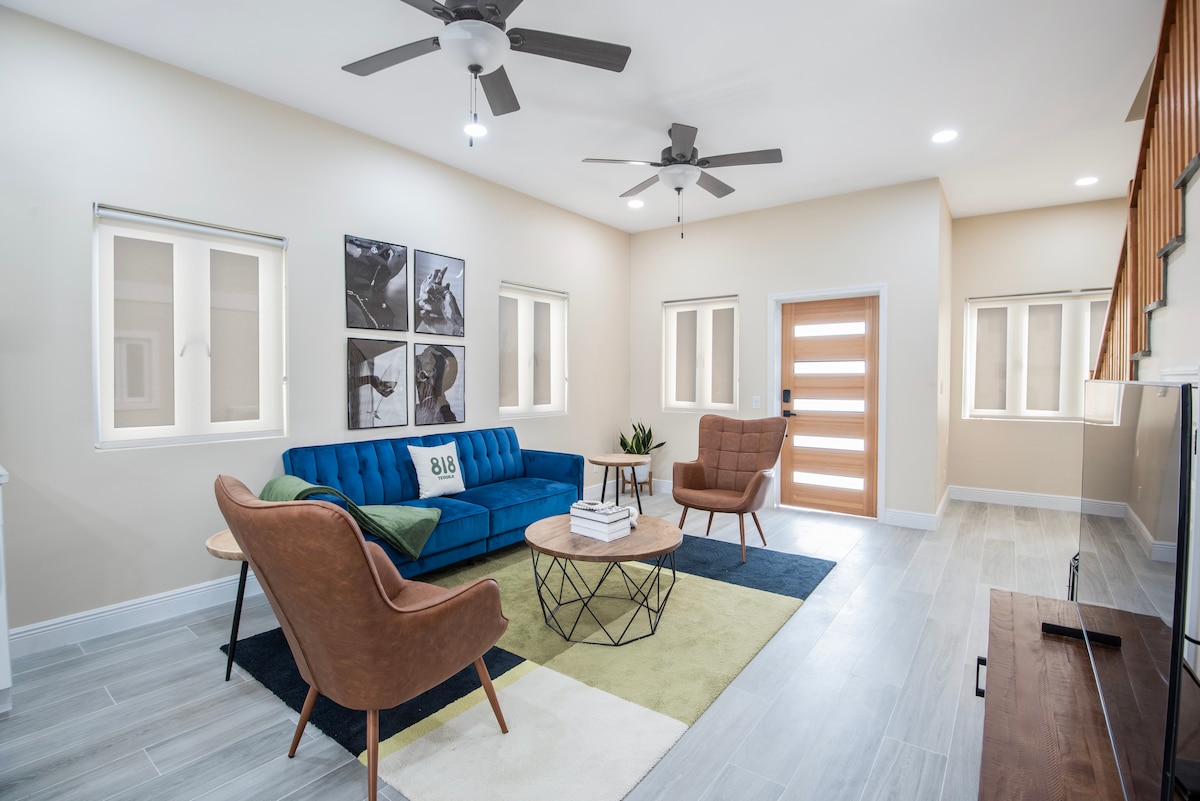 A spacious living area features a blue sofa, two brown armchairs, and a geometric coffee table. Light-colored walls complement the natural light from multiple windows. A stylish wooden door and modern ceiling fans enhance the inviting ambiance of the space.