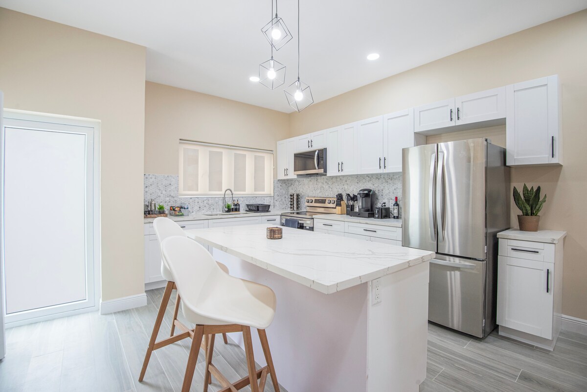 A modern kitchen is showcased with white cabinetry and a marble countertop. A large stainless steel refrigerator is present, along with a sleek stove and microwave. Bar stools are positioned at the island, complemented by soft overhead lighting.