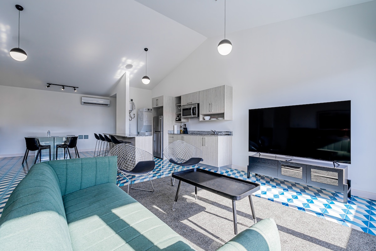 An open-concept living area features a modern design with a green sectional sofa, a coffee table, and a large television. The kitchen is equipped with stainless steel appliances and light cabinetry. A dining area with black chairs is visible in the background, alongside a geometric tiled floor.
