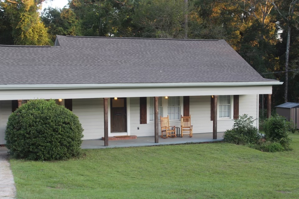 The exterior of the house features a welcoming porch with two wooden rocking chairs. Lush green grass surrounds the front yard, which is framed by trees in the background. The structure has a gabled roof and light-colored siding, contributing to a charming entrance.