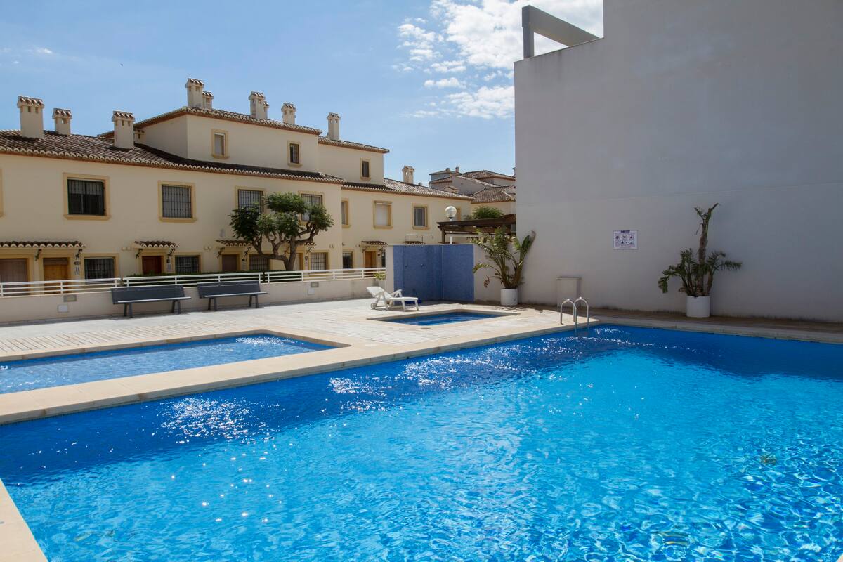 The inviting pool area features a bright blue swimming pool surrounded by light stone decking. Additional smaller pools are positioned for relaxation. Well-maintained greenery adds a touch of nature, while nearby residential buildings provide a sense of community.
