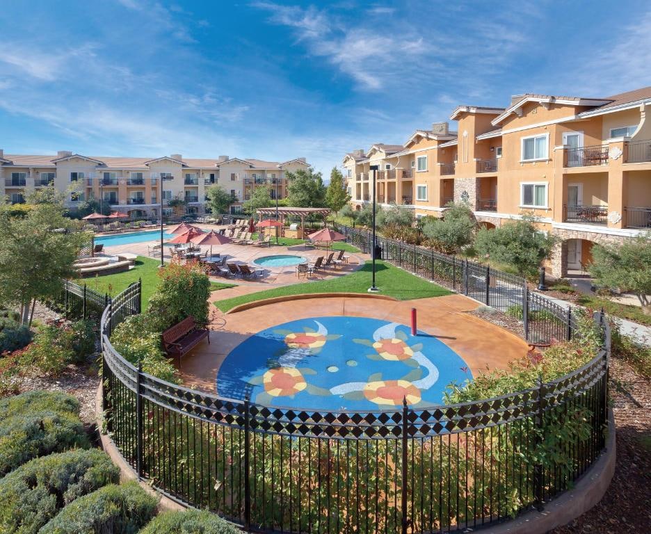 An outdoor area features a colorful splash pad surrounded by well-maintained gardens. The spacious pool area is visible, with lounge chairs arranged for relaxation. The buildings in the background are multi-story, offering balconies for guests to enjoy views of the grounds.
