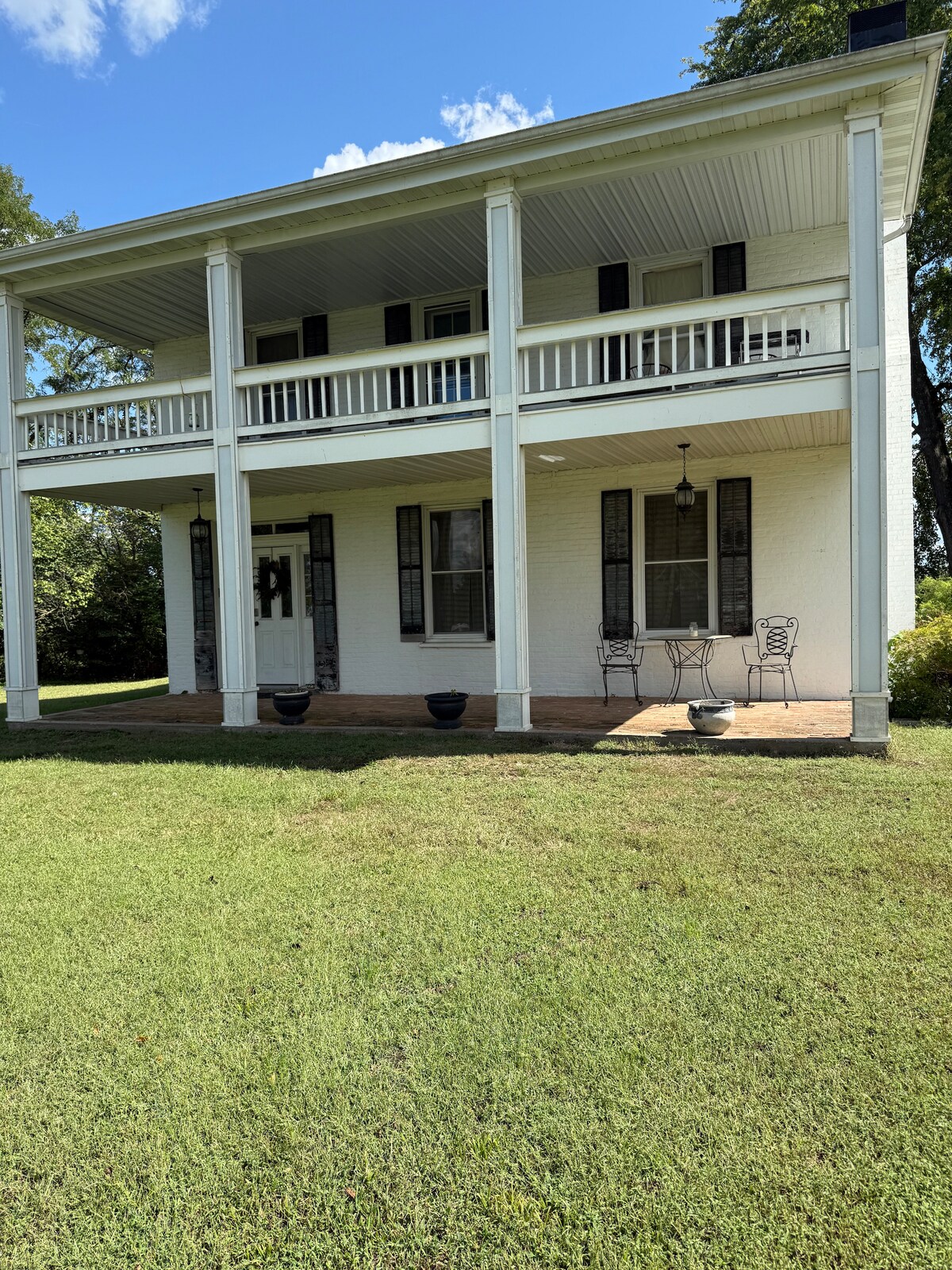 A two-story house features a spacious porch with white columns, two sets of black chairs, and small tables. The green lawn surrounds the exterior, while large windows and doors on the upper and lower levels allow natural light to enter the home.