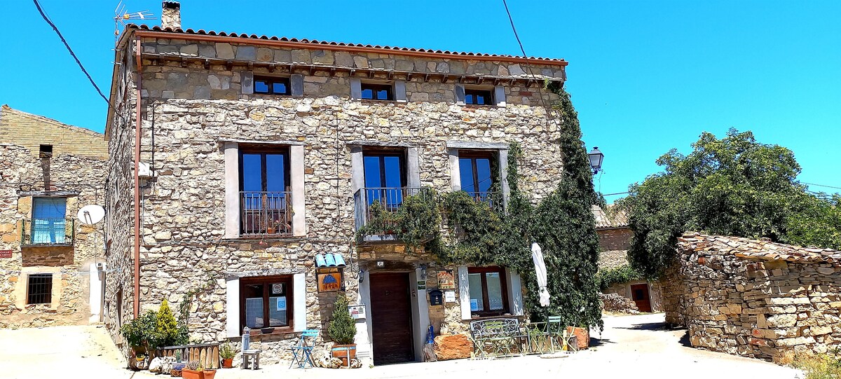 A charming stone house features a rustic facade, with multiple large windows framed by greenery. A wooden door is centrally positioned, while a small balcony on the second floor offers a view of the surrounding area. Outdoor seating is visible at the entrance.