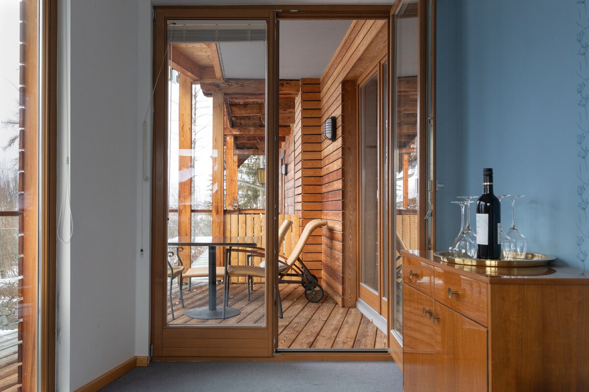 A modern living area features large glass doors opening to a wooden terrace. The terrace includes a pair of comfortable chairs and a small table. A wooden cabinet is visible in the corner, holding a bottle of wine and a tray.