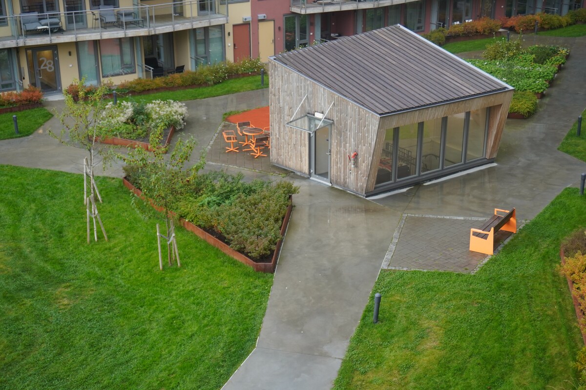 An outdoor area is highlighted by a contemporary wooden structure featuring large glass windows. Surrounding greenery includes landscaped gardens and seating areas, along with paths made of smooth concrete and natural grass. The setting provides a serene environment for relaxation.