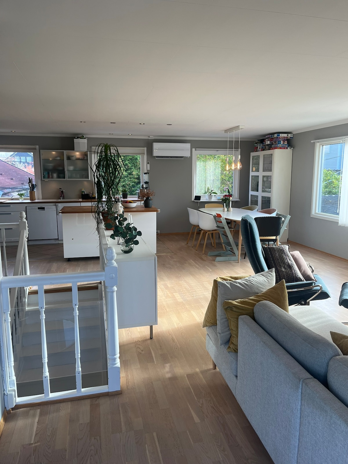 An open kitchen and living area is depicted, featuring modern appliances and a dining table surrounded by chairs. Natural light enters through large windows, enhancing the airy feel. A comfortable sofa is positioned in the foreground, and a plant adds a touch of greenery to the space.
