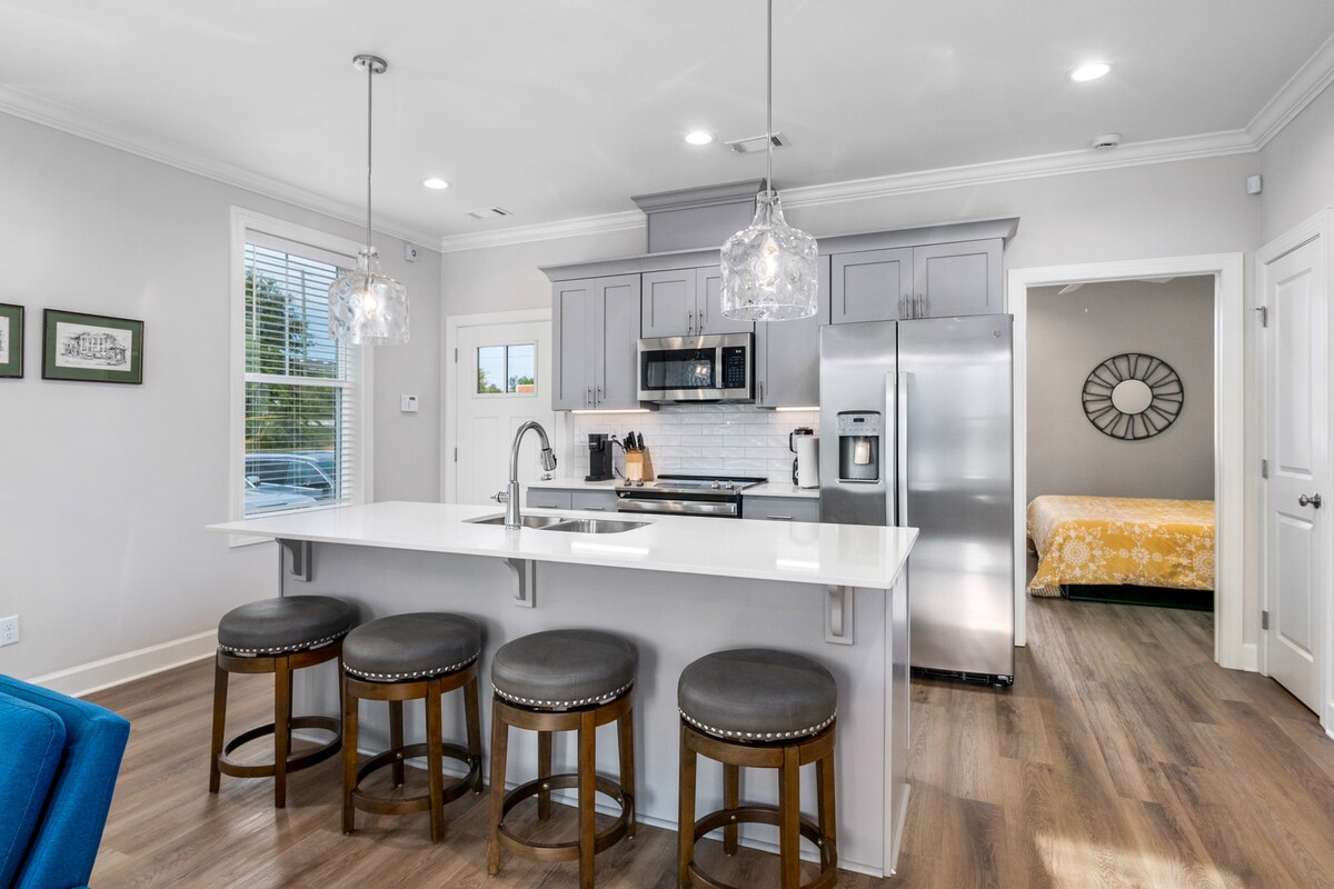 A modern kitchen features sleek gray cabinetry and stainless-steel appliances. The kitchen island is highlighted by four bar stools with brown leather upholstery. Pendant lights hang above the island, enhancing the bright and open atmosphere. A doorway leads to a bedroom area in the background.