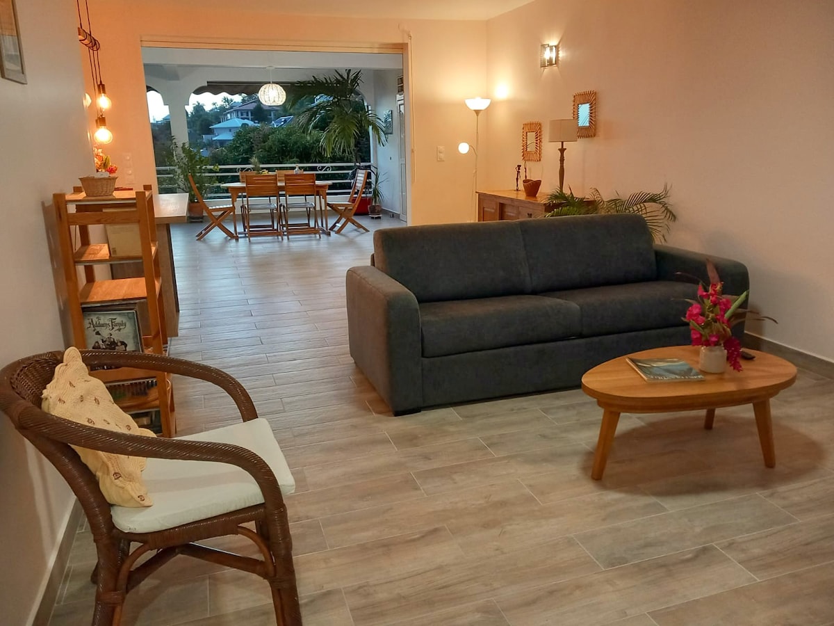 An open living area features a gray sofa and a wooden coffee table. A rattan chair adds a touch of warmth. Behind, a dining space is visible with wooden furniture, and large sliding doors allow natural light to fill the room, providing a view of the outdoor terrace.