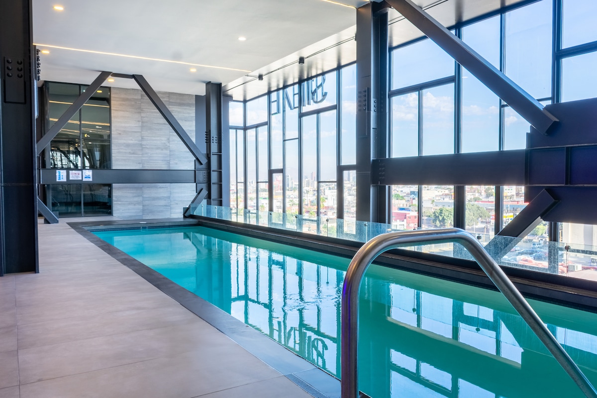 A bright indoor pool area features floor-to-ceiling windows with city views. The minimalist design includes sleek metal supports, and the water reflects natural light. A staircase leads into the pool, surrounded by tiled flooring, enhancing a spacious and open environment.