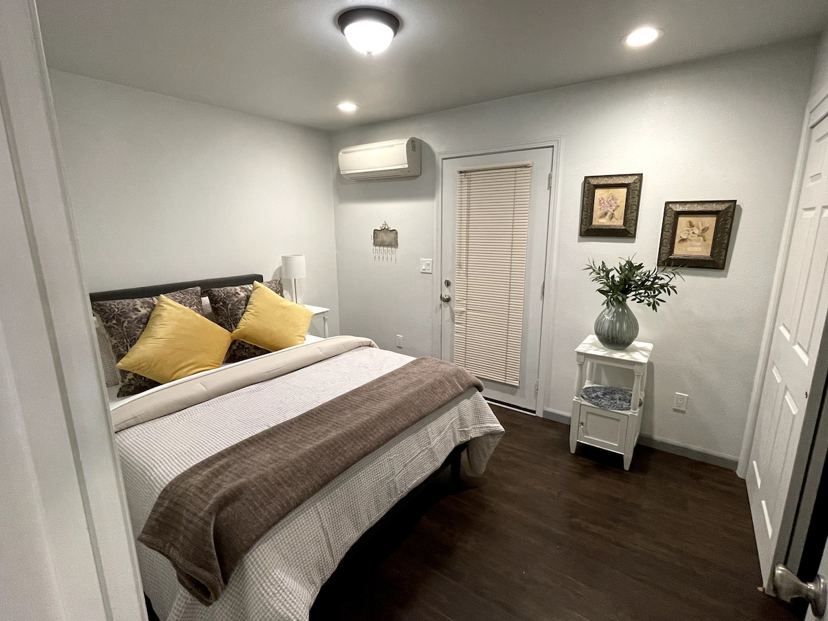 A well-furnished bedroom features a spacious bed adorned with neutral linens and decorative yellow pillows. A small nightstand with a lamp is present beside the bed. Natural light enters through a sliding door, which opens to an outdoor area. Framed art pieces and a plant add subtle decor.