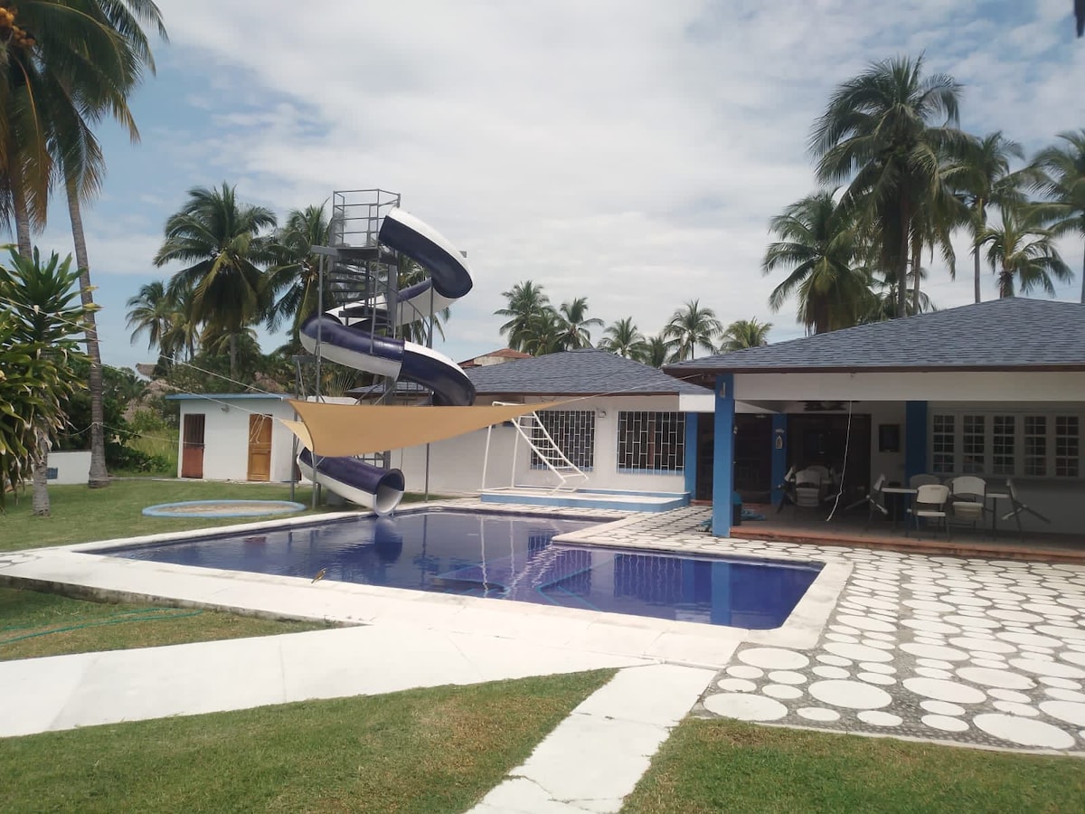 An outdoor area features a swimming pool surrounded by palm trees, with a waterslide positioned nearby. A spacious patio includes a shaded seating area, offering relaxation options. The layout showcases a blend of grass and tiled surfaces, creating an inviting space for leisure.