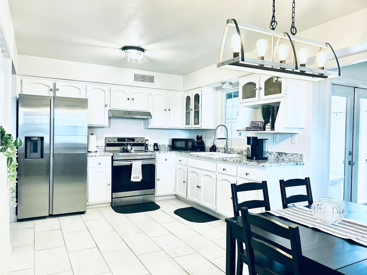 An inviting kitchen features modern stainless steel appliances and ample counter space, complemented by white cabinetry. A dining table with dark wood chairs is present beneath a stylish light fixture, creating a welcoming area for meals or gatherings. Natural light is facilitated through large windows.