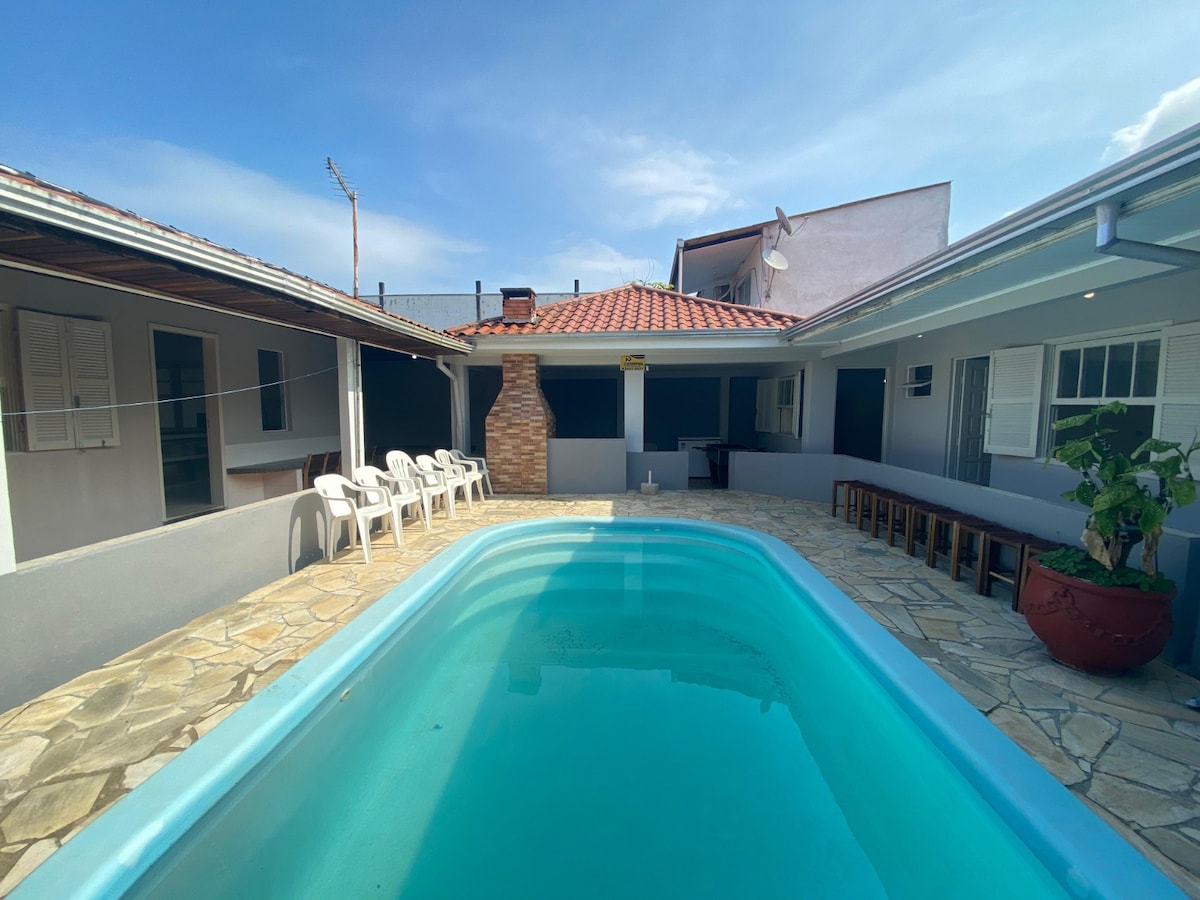 A crystal-clear swimming pool is centered in a stone-paved courtyard, bordered by comfortable seating arrangements. The rustic barbecue area is visible beyond the pool, and the surrounding spaces are enclosed with windows, allowing ample natural light.