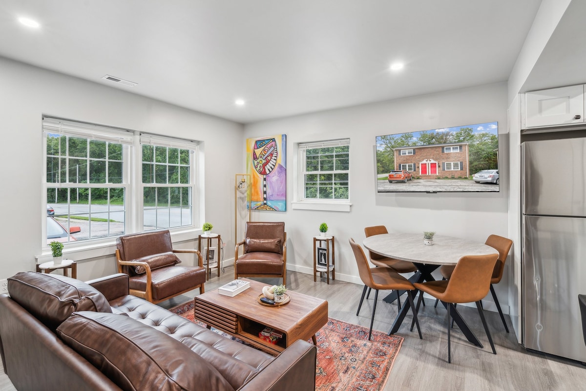 A modern living area features brown leather seating and a wooden coffee table. Natural light enters through large windows, illuminating the space. A round dining table with tan chairs is positioned nearby, and a flat-screen TV displays an outside view.