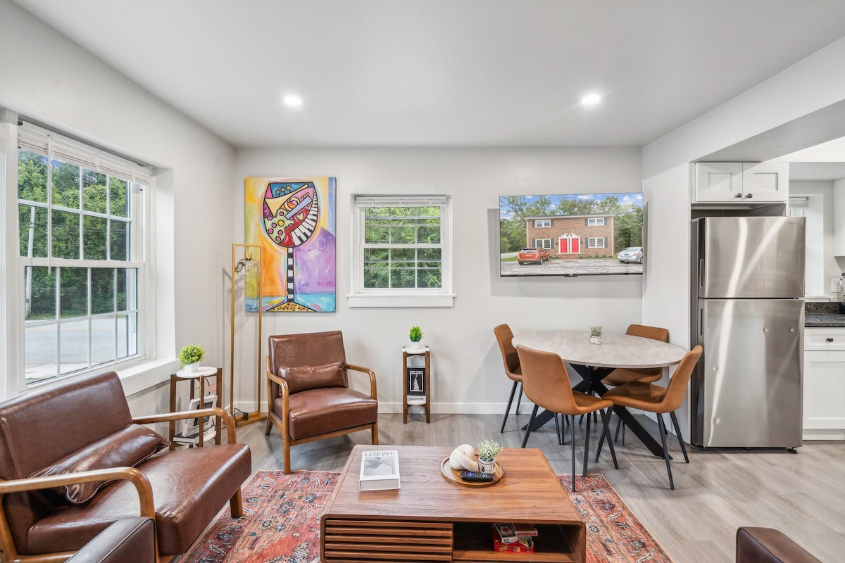 A modern living space features a round dining table surrounded by four chairs, paired with a wooden coffee table. A cozy seating area includes two leather armchairs and a vibrant art piece on the wall. Natural light enters through large windows, highlighting the space.