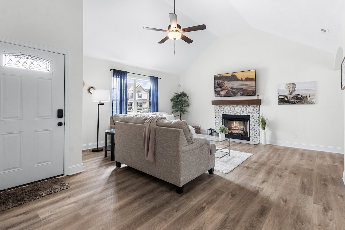 A light-filled living area features a comfortable sofa facing a stylish fireplace, framed by decorative tiles. A ceiling fan and large windows enhance the airy feel, while potted greenery adds a touch of nature to the inviting space.