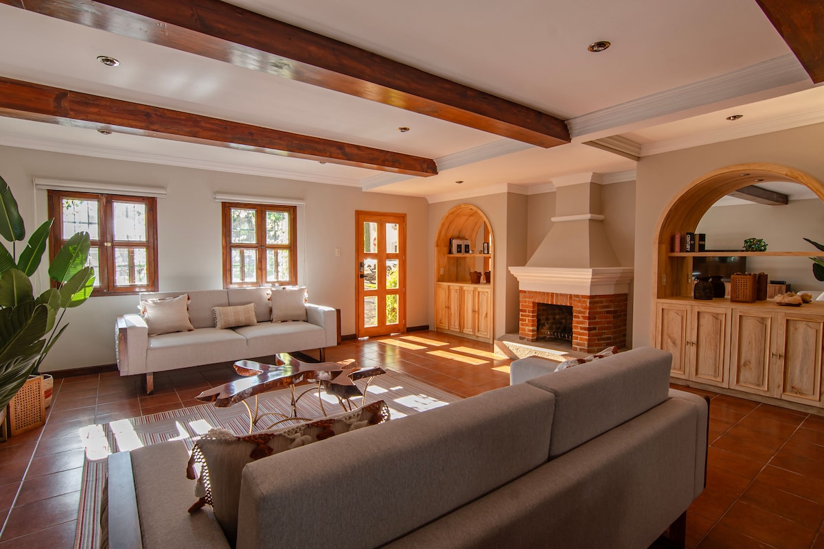 A spacious living area is presented with two sofas arranged around a coffee table. Large windows allow ample natural light to fill the room, accentuating the warm wooden beams overhead. A traditional fireplace is featured, along with built-in shelving displaying decor elements.