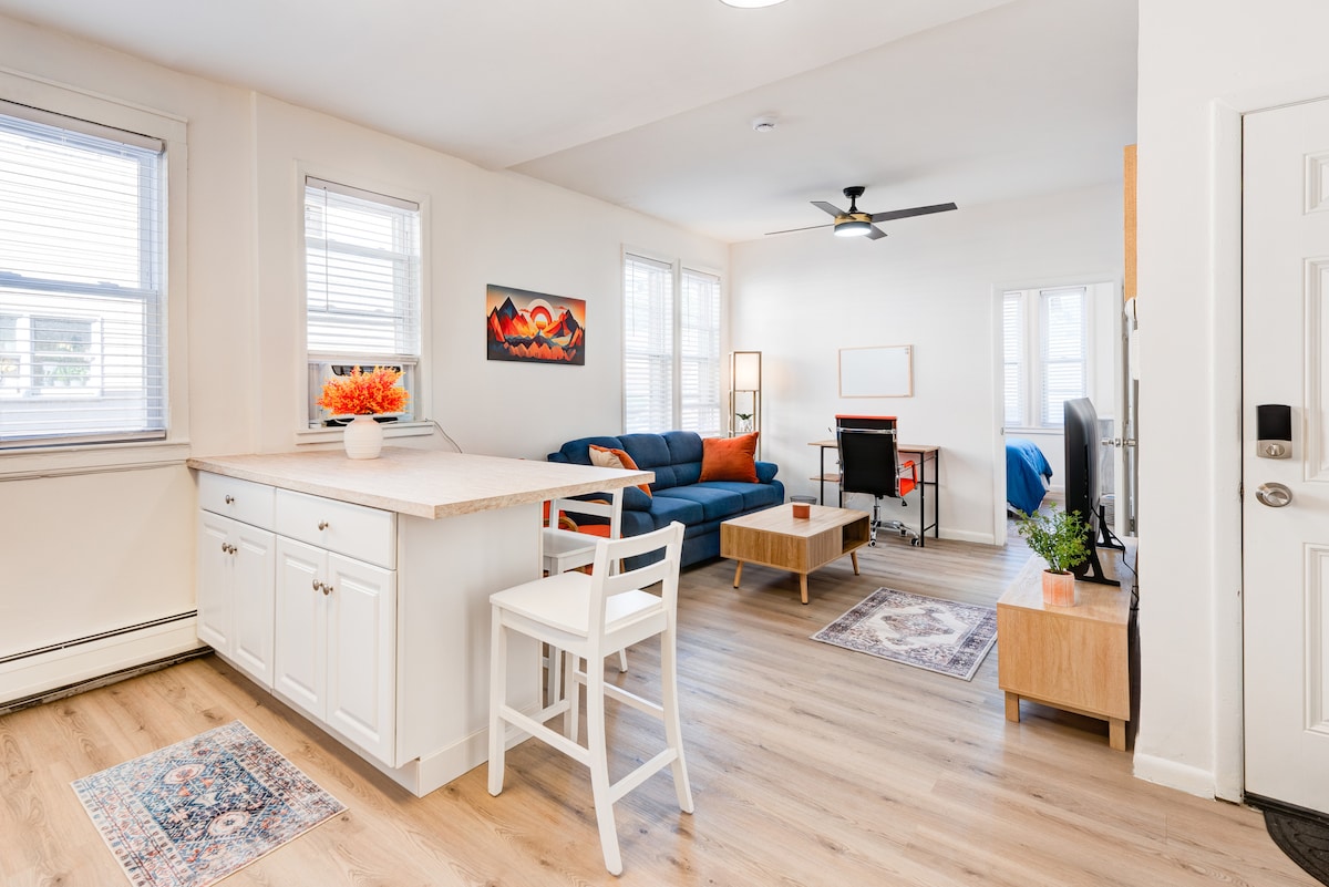 A bright living space features a comfortable blue sofa, complemented by a wooden coffee table. Natural light enters through four windows, enhancing the warmth of the light-colored walls and wooden flooring. A small dining area with a white chair and a simple kitchen island can be seen.