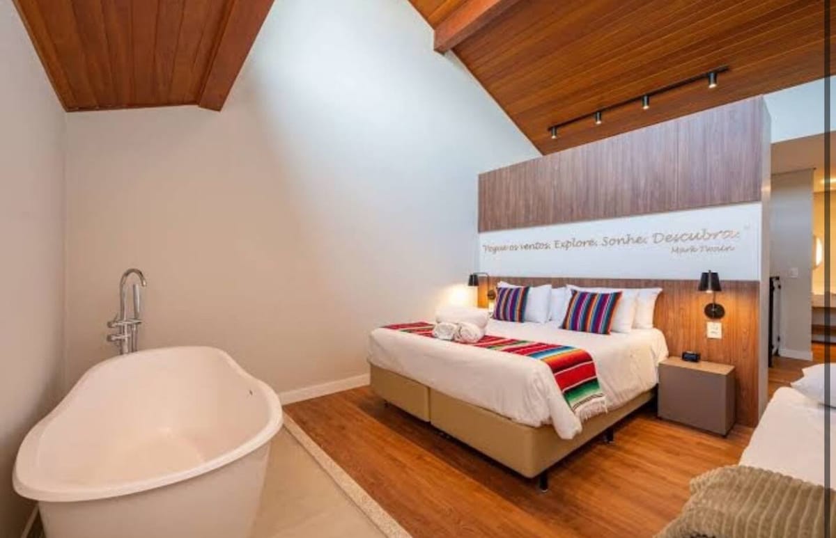 A spacious bedroom features a large bed with colorful decorative pillows and a minimalist clawfoot bathtub nearby. Soft natural light filters in through overhead windows, illuminating the warm wood accents of the sloped ceiling and enhancing the room's elegant design.