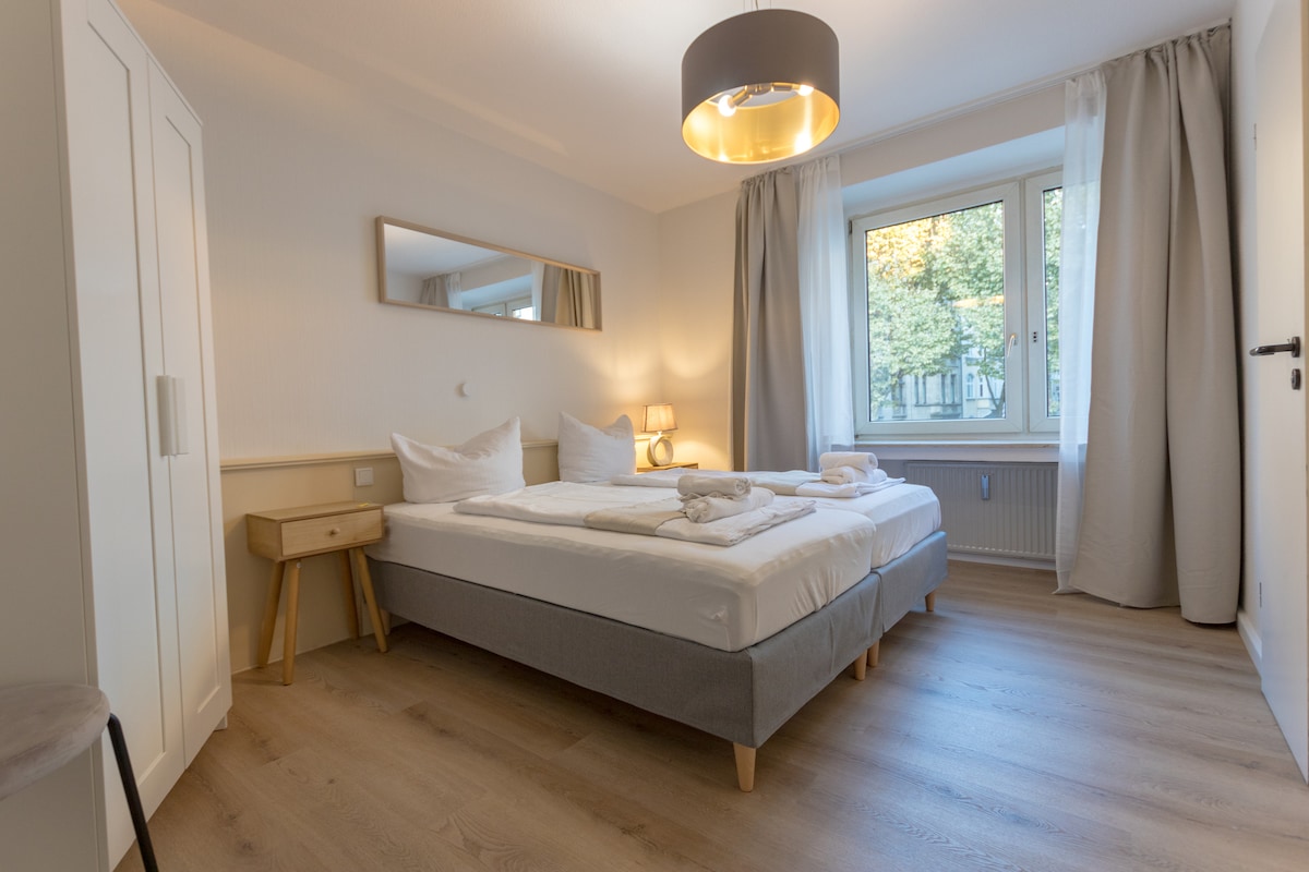 A well-lit bedroom features two spacious beds with crisp linens, complemented by soft towels neatly arranged on top. A wooden nightstand and a modern lamp provide practical lighting. Large windows invite natural light, while sheer curtains soften the view.