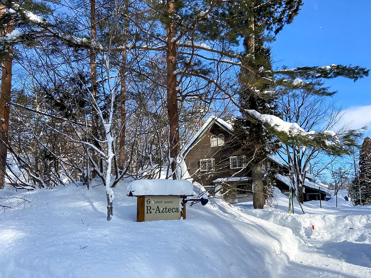 A snow-covered entrance to the R-Azteca pension is framed by tall trees, with a sign partially visible in the foreground. The wooden building is nestled within the snowy landscape, showcasing a rustic charm against a clear blue sky.