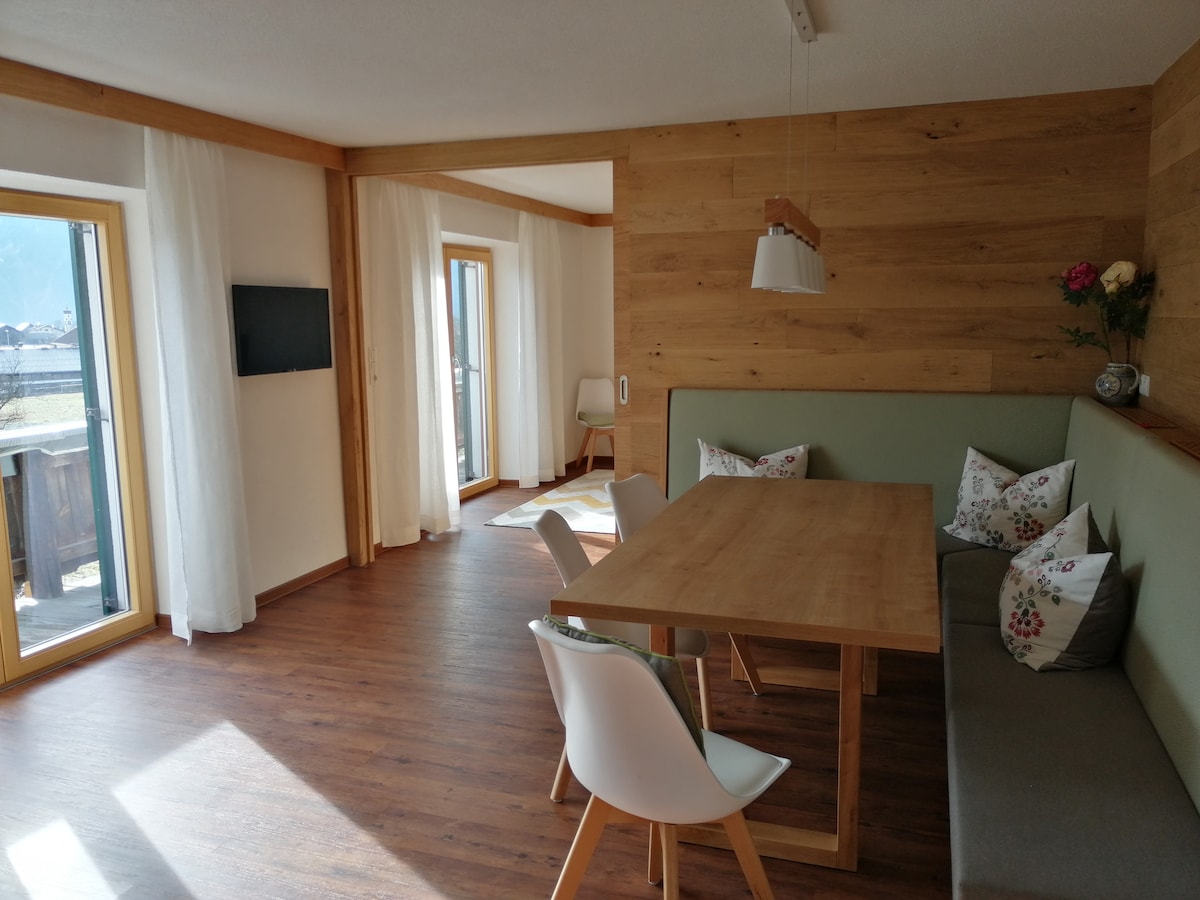 A bright and airy dining area features a wooden table surrounded by four chairs. Natural light filters through large windows with sheer curtains, illuminating a cozy seating nook with decorative cushions. A wall-mounted television is visible in the background.