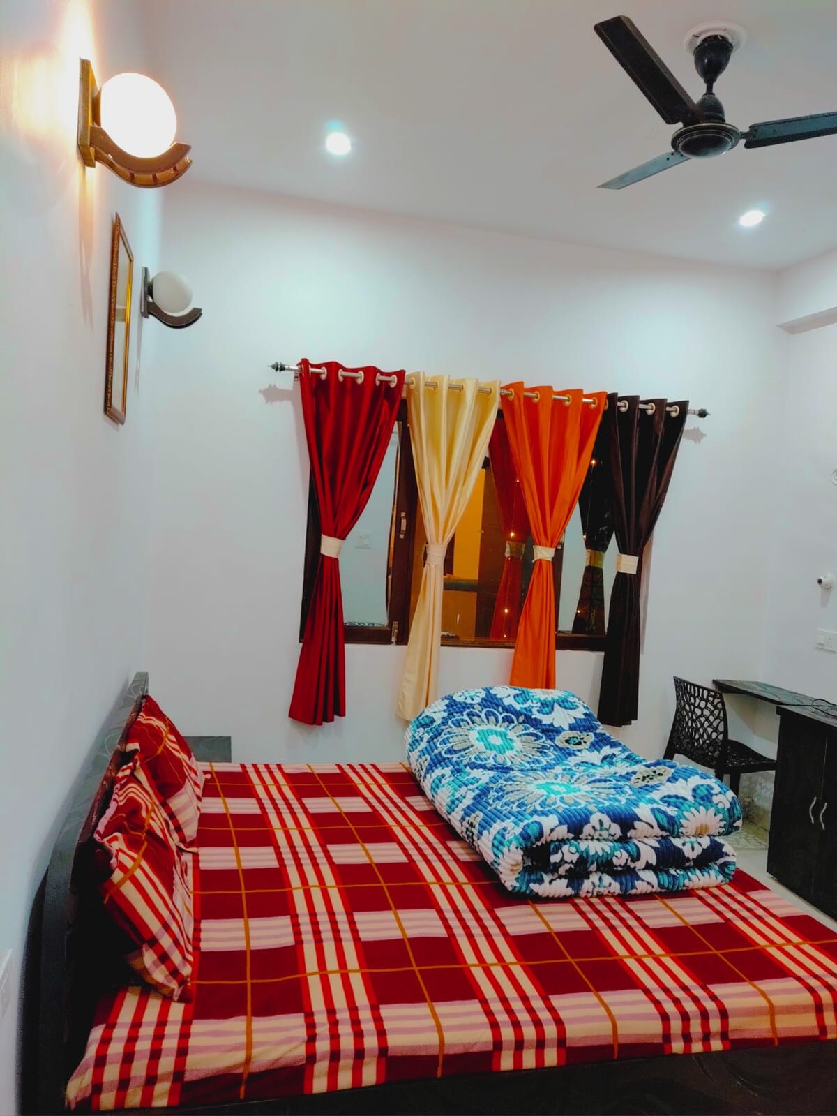 A cozy sleeping area is organized with a spacious bed, layered with a patterned blanket and decorative throw pillows. Colorful curtains in red, orange, and brown frame the window, which allows soft light to illuminate the room. A desk with a chair is positioned nearby.