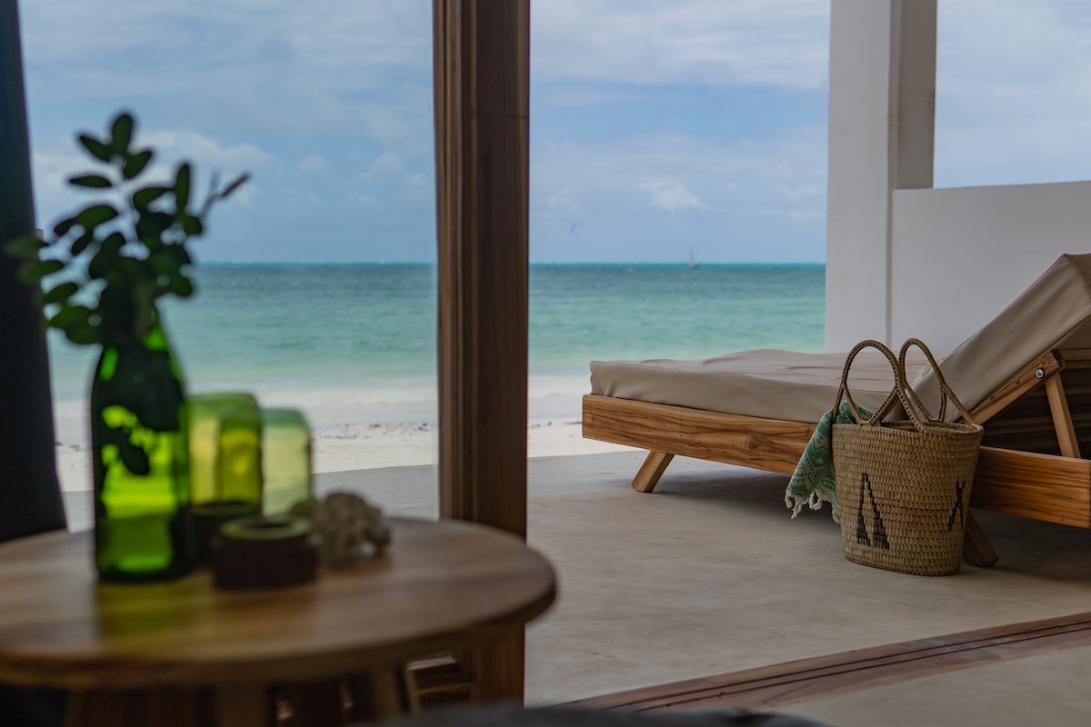 A wooden lounge chair is positioned to overlook a sandy beach and calm turquoise waters. Green glass vases and a woven basket are set on a nearby table, complementing the relaxed coastal ambiance. Soft natural light fills the space through open doors.