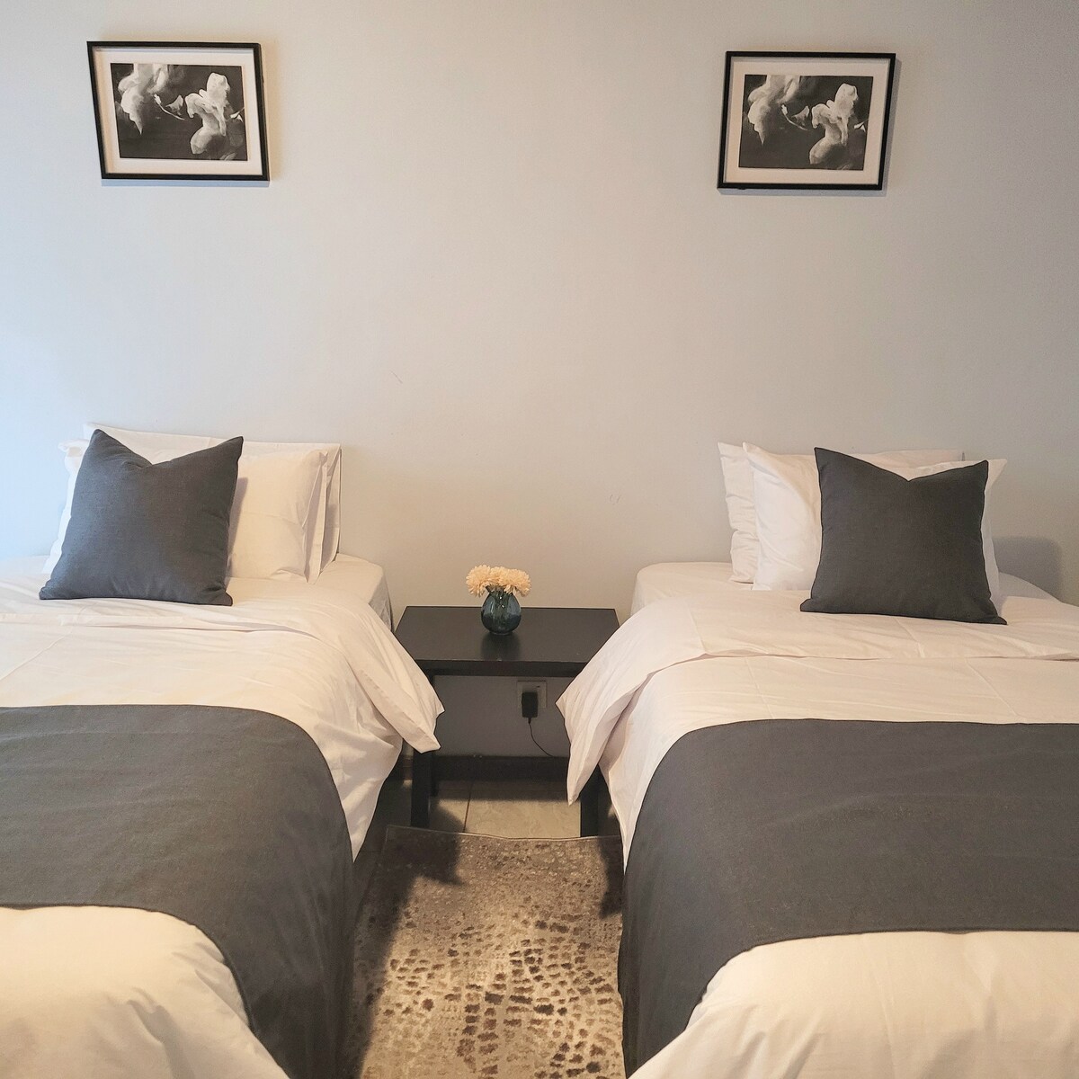 Two twin beds are positioned side by side, each adorned with gray throw pillows. A small bedside table, holding a vase with flowers, is placed between the beds. Minimalistic wall art complements the soft color scheme, creating a serene sleeping area.