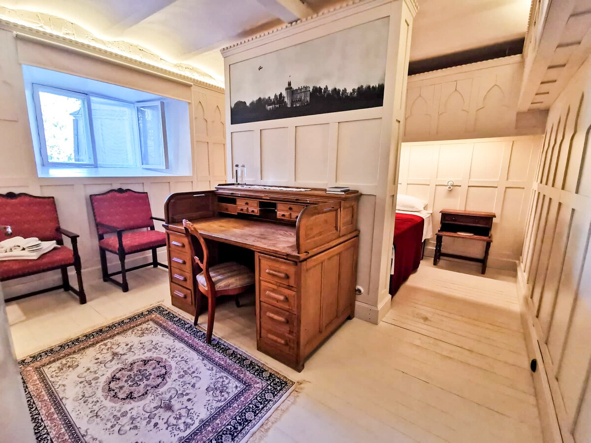 A warm, inviting room features a wooden desk with multiple drawers and two comfortable chairs placed nearby. A patterned area rug adds character to the floor. A small bed is positioned in the corner, with natural light filtering through a window.