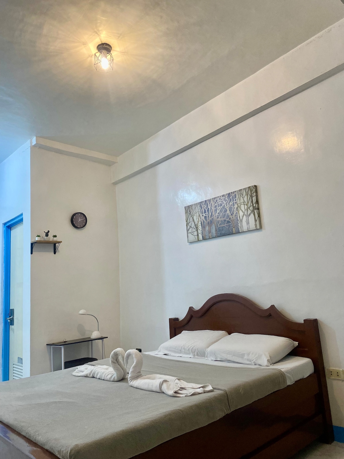 A cozy sleeping area features a wooden bed with neatly arranged pillows and bath towels. A simple bedside table holds a lamp, while a wall art piece depicting trees adds a touch of nature. Soft light illuminates the space from a ceiling fixture.
