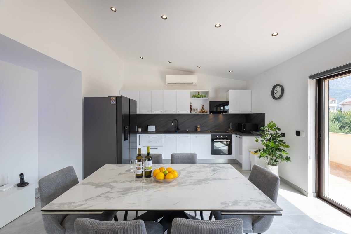 A modern kitchen and dining area features a large marble table surrounded by gray chairs. The kitchen is equipped with white cabinetry, a dark countertop, and various appliances. Natural light fills the space through adjacent sliding glass doors, offering a view of the balcony.