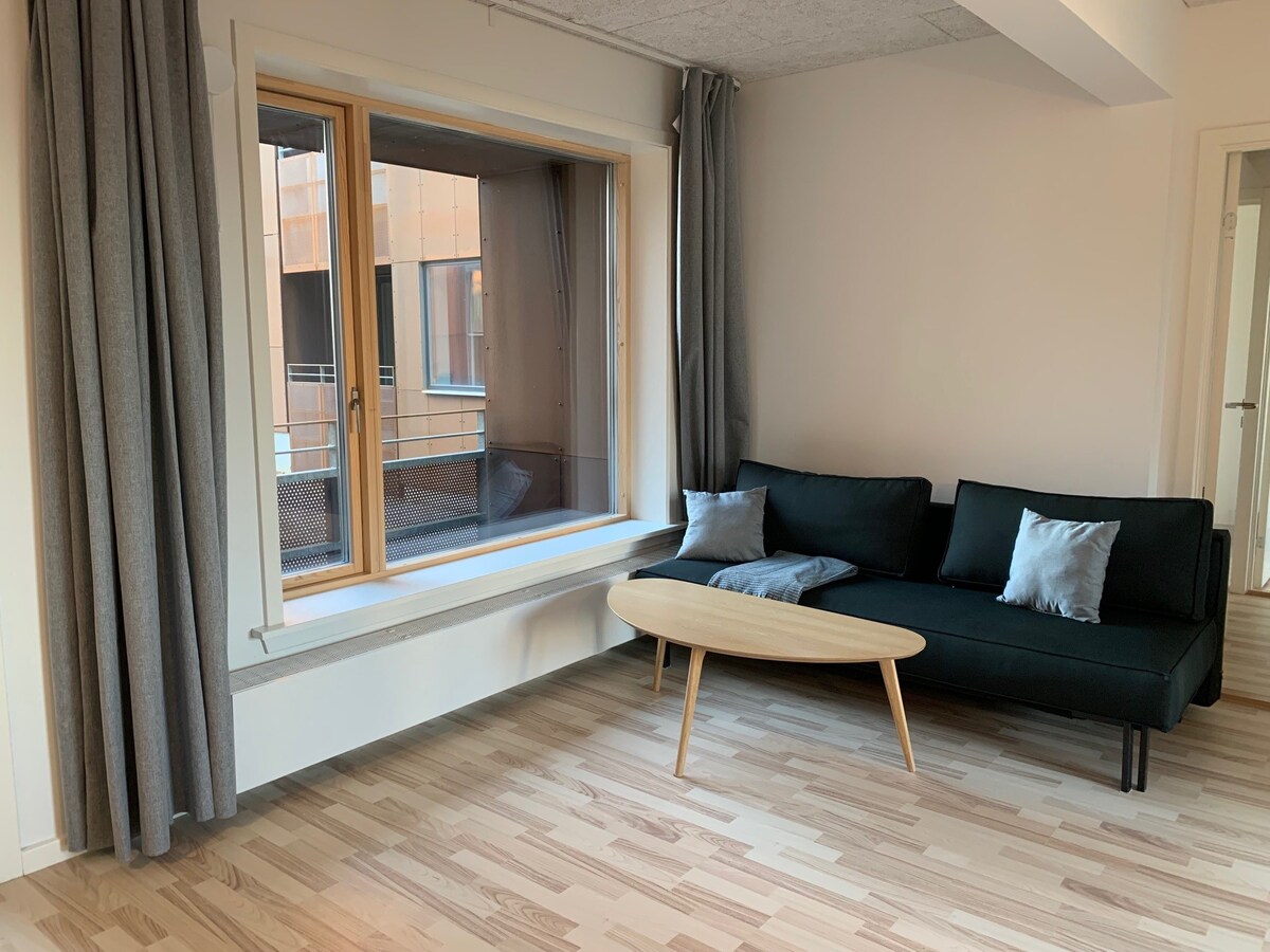 A spacious living area features a comfortable dark sofa accented by two light grey cushions. A wooden coffee table is positioned in front, while a large window allows natural light to illuminate the space, providing a view of the courtyard outside.
