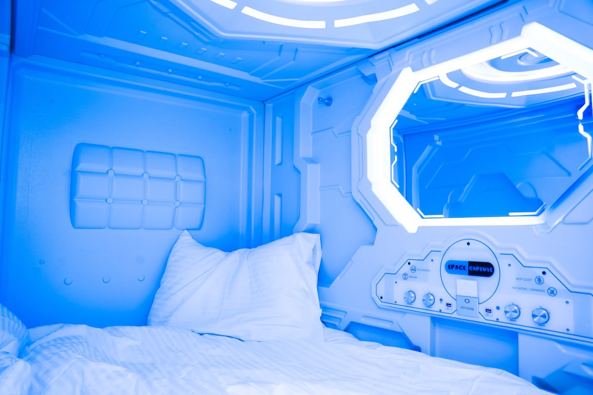 An enclosed sleeping area features soft white bedding arranged atop a spacious mattress. Sleek blue lighting casts a calming glow, enhancing the futuristic design. A reflective surface on the opposite wall adds depth, while controls are positioned conveniently for ease of access.