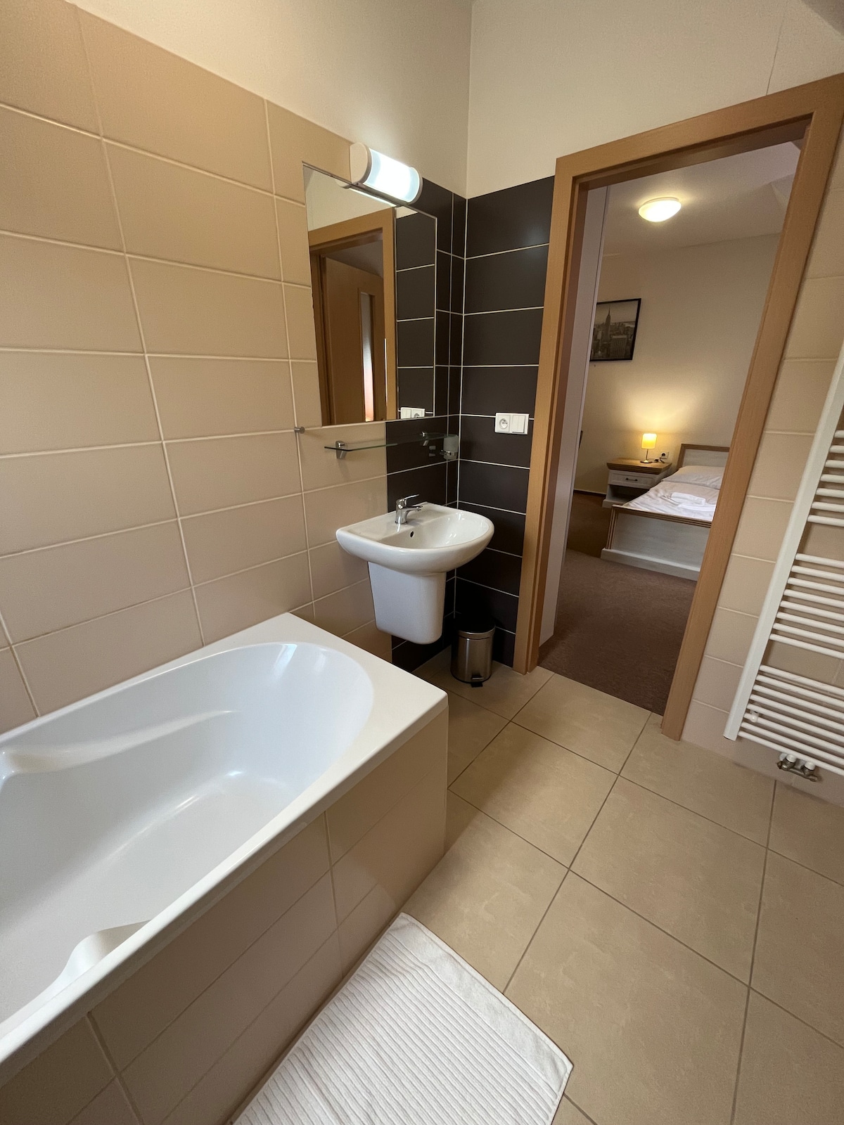A modern bathroom features a white bathtub and a compact sink mounted on a light-colored wall. A full-length mirror is positioned above the sink. The entrance opens towards a softly lit bedroom, visible in the background, with a glimpse of a cozy bed.