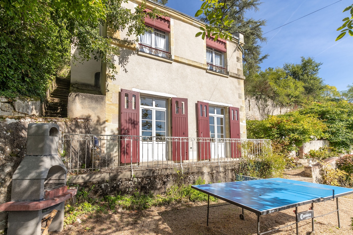 The exterior of a semi-troglodyte house showcases large windows framed by burgundy shutters. A stone barbecue is positioned beside a blue ping-pong table, inviting outdoor activities. Lush greenery surrounds the structure, providing a serene setting with steps leading to the entryway.