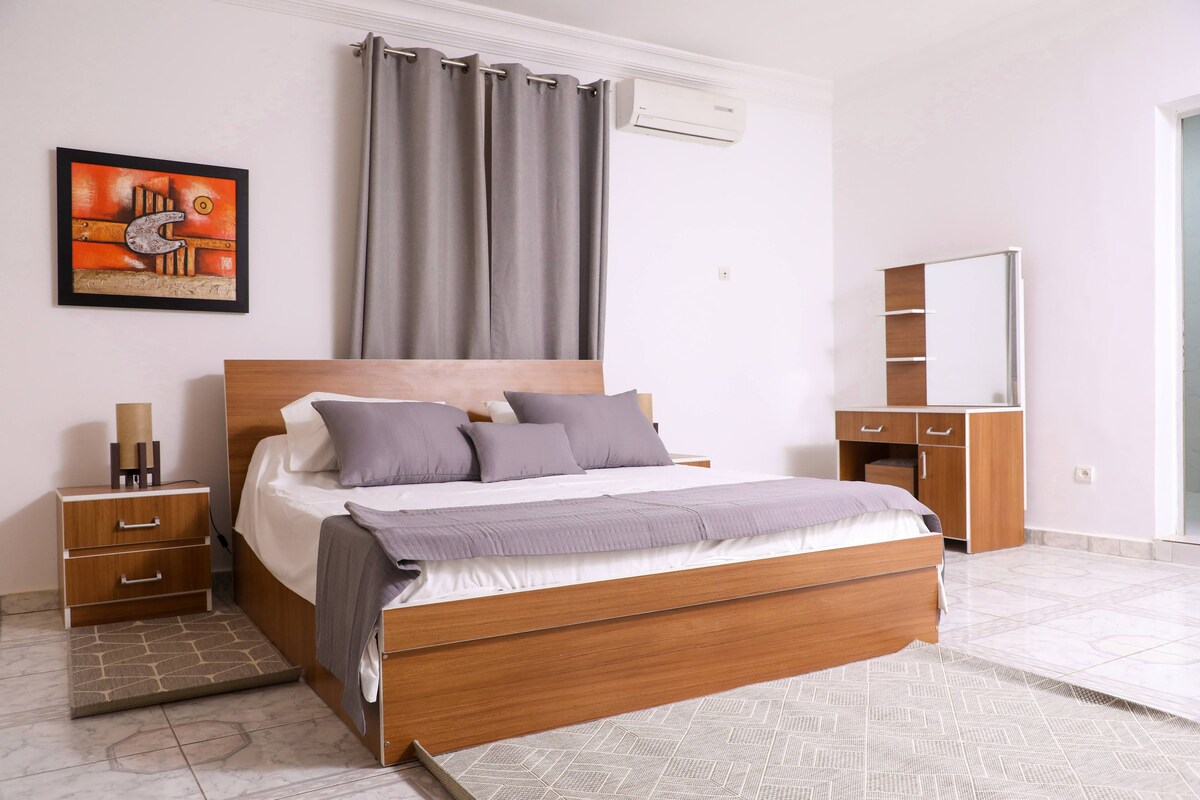 A spacious bedroom is furnished with a large wooden bed featuring layered bedding in neutral tones. Gray curtains frame a window, while a dresser and side tables complement the decor. A colorful piece of wall art adds a touch of character to the minimalist design.